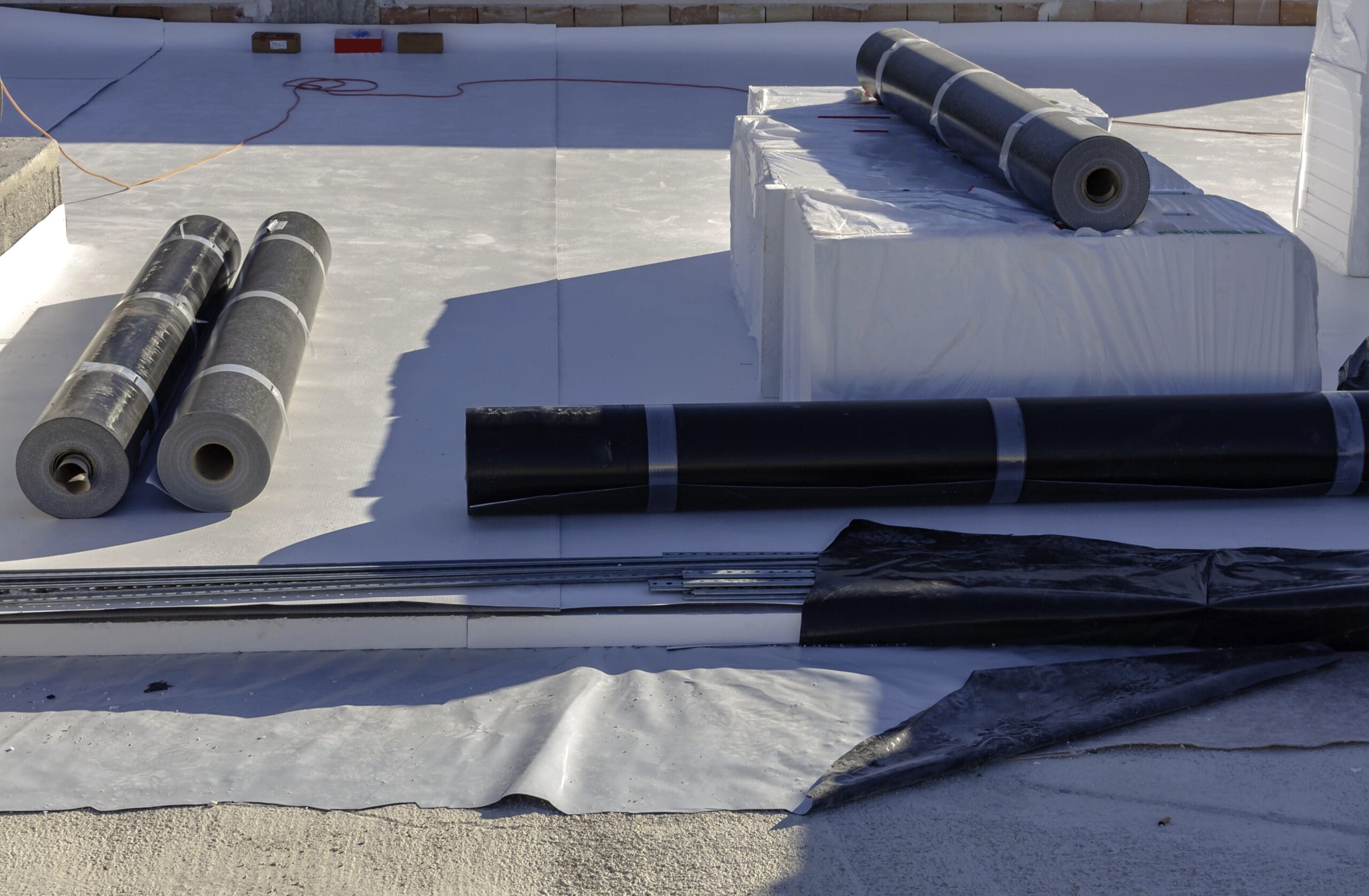 Three rolls of black waterproofing material lie on a flat roof, alongside additional rolls wrapped in white plastic. Metal rods and black sheets hint at ongoing construction or renovation work for new flat roofs or upcoming chimney works.